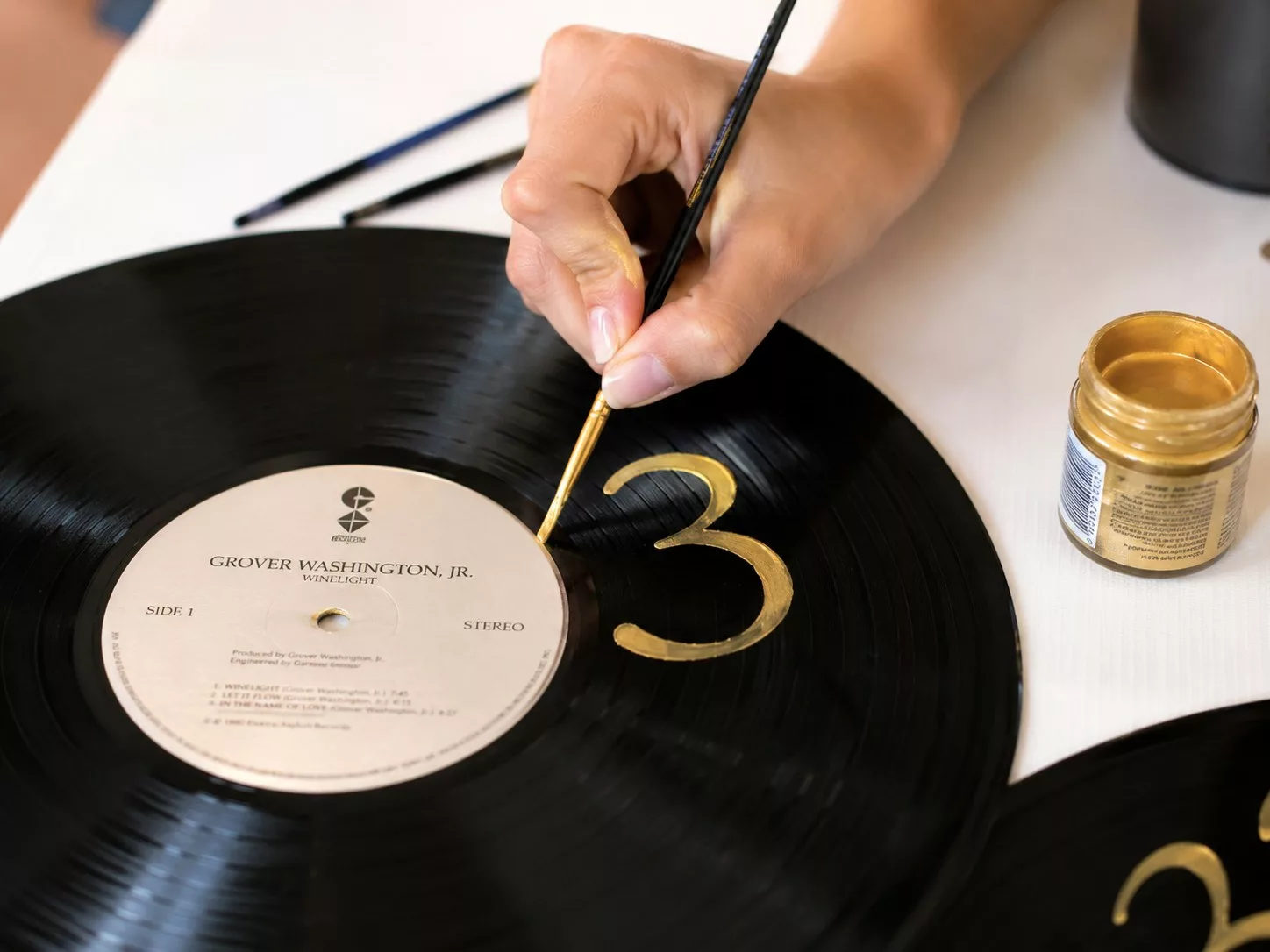 Main peignant un numéro de table doré sur un disque vinyle, DIY mariage musique