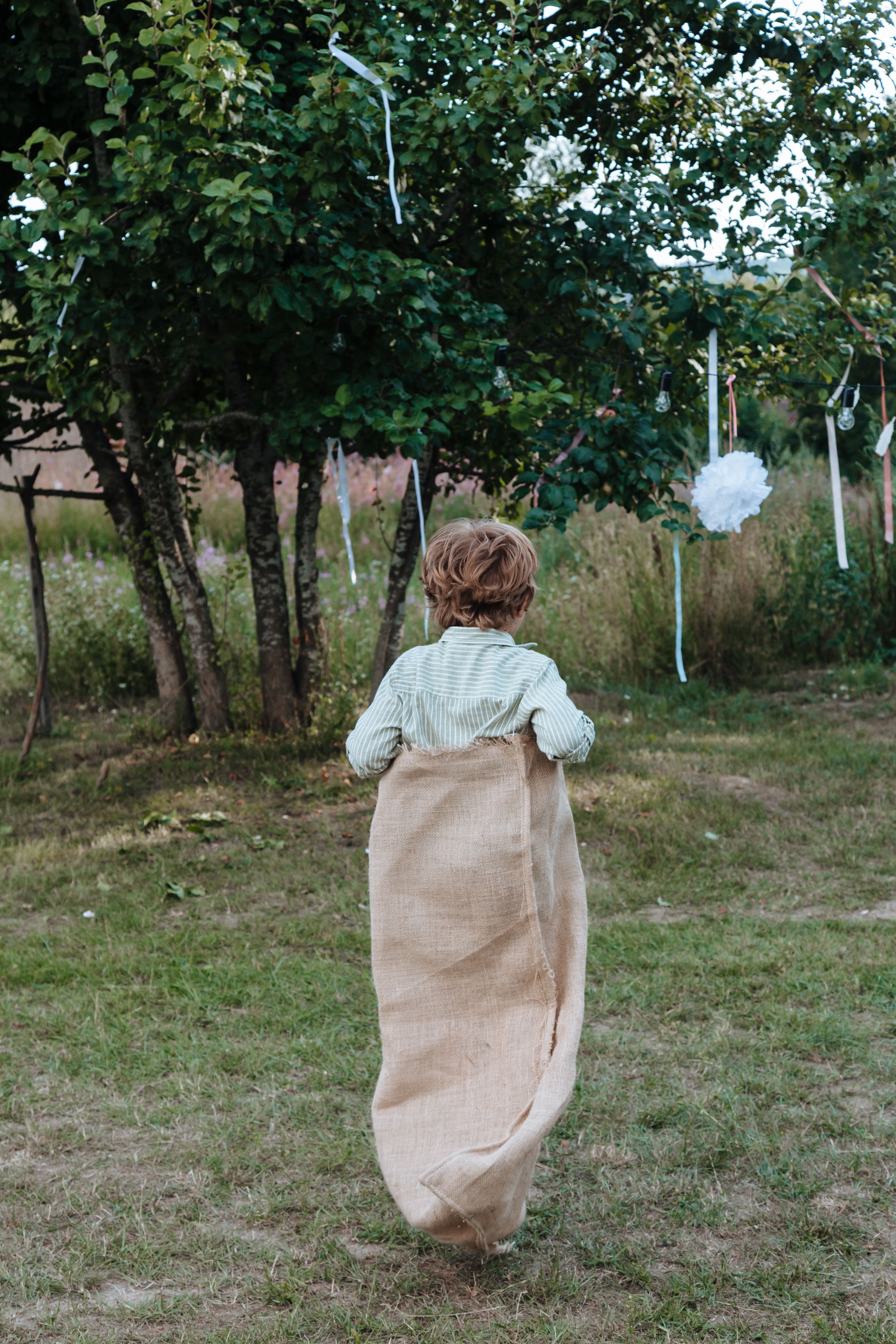 Enfant en course dans un sac de jute sur une pelouse decoree de guirlandes et de pompons blancs de mariage